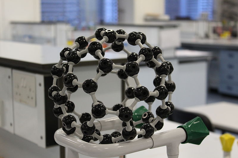 A photograph of a ball and stick model of fullerene sitting on holder on a lab bench is pictured. Each carbon atom is bonded to three other carbon atoms, which results in the formation of a ball like structure.