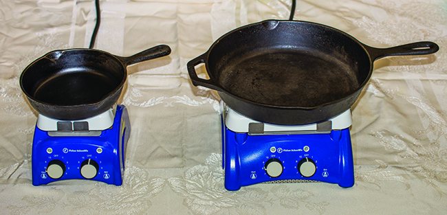 Two black metal frying pans sitting on a flat surface are pictured. The left pan is about half the size of the right pan.
