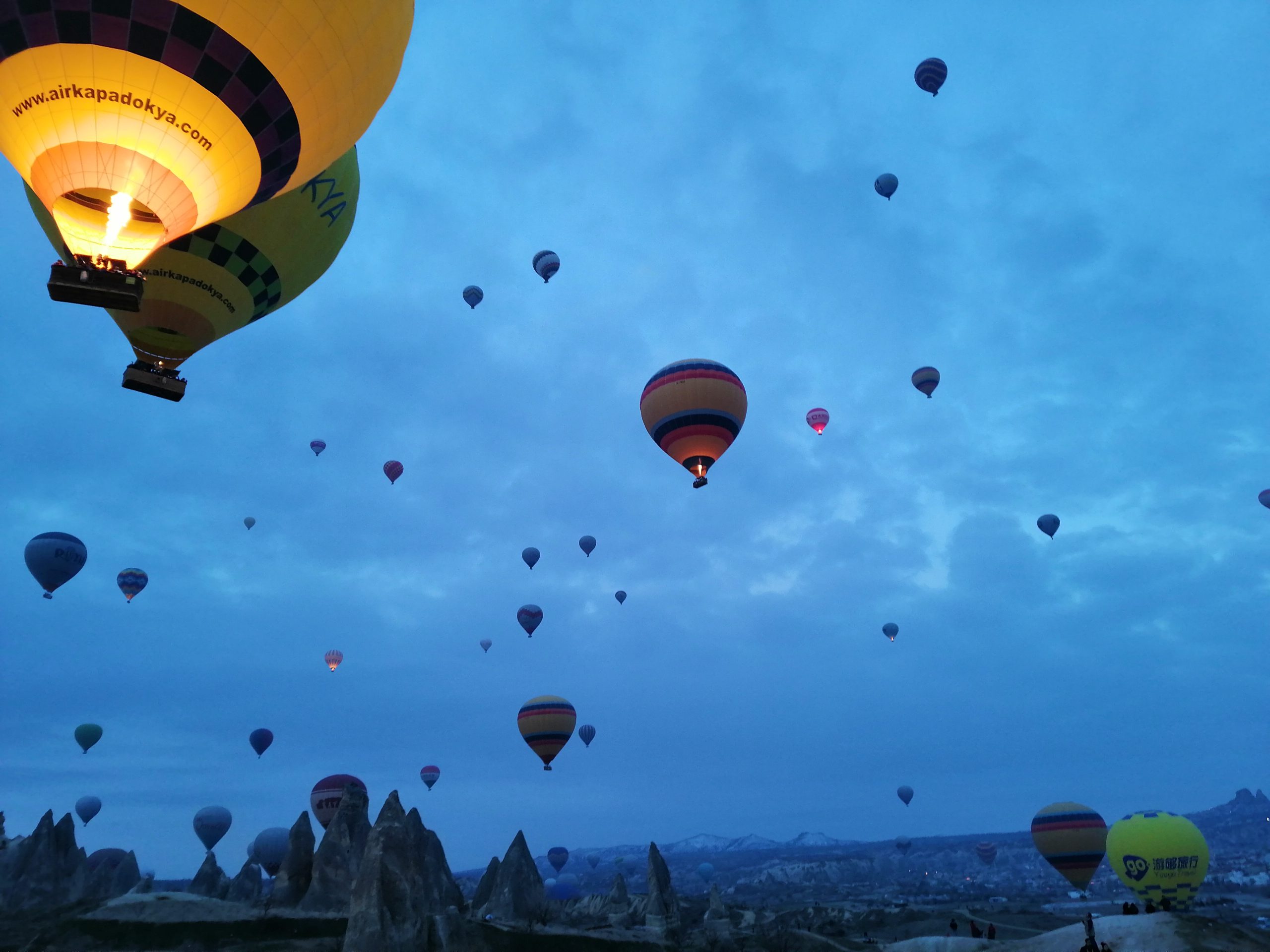 A photograph shows about thirty colorful hot air balloons in the air at various distances against a blue twilight sky.