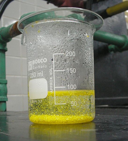 Inside a 250 mL beaker, yellow hexagonal crystals are forming in aqueous solution. Solids have also settled at the bottom of the beaker.