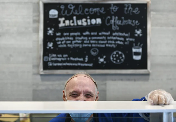 A white older man wearing a mask and clear gloves is smiling at the camera with one hand resting on the counter of the Inclusion Coffeehouse.