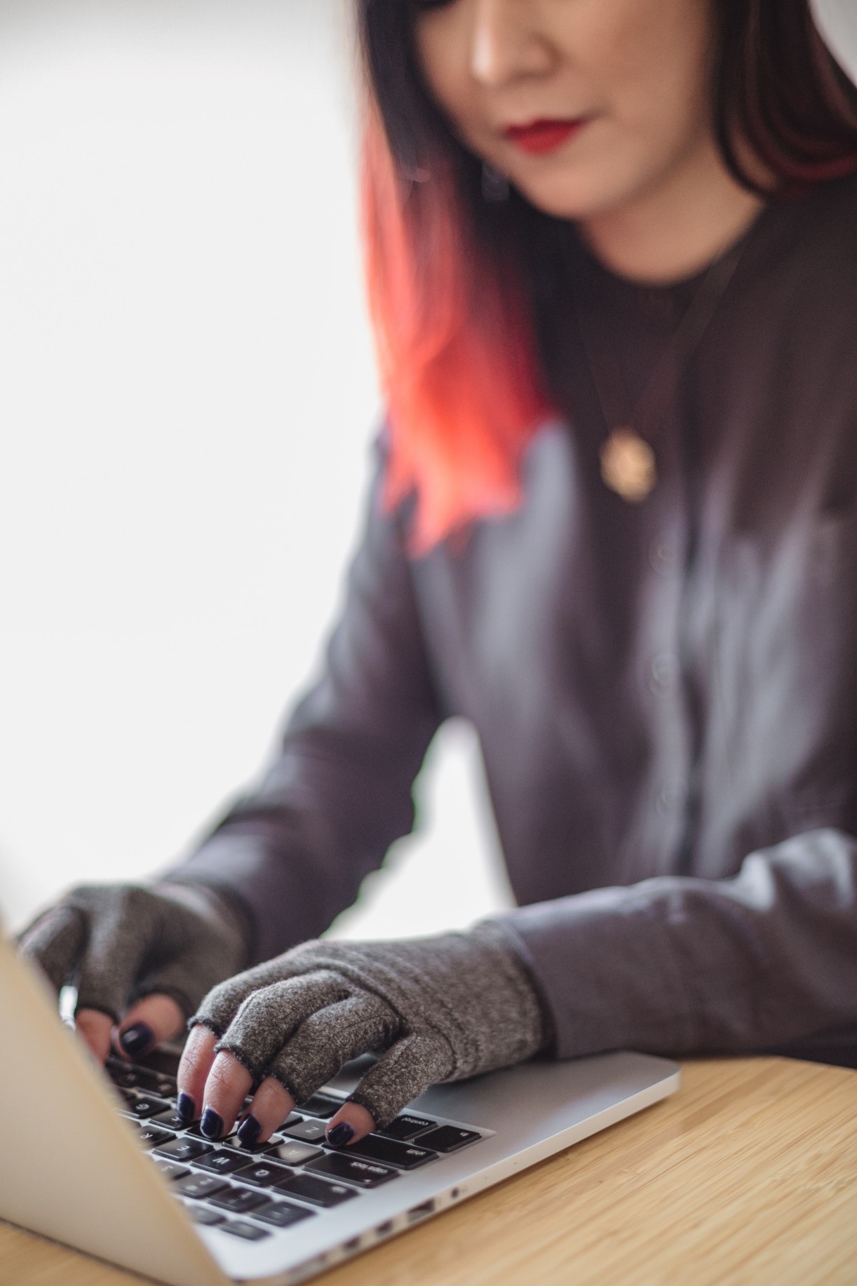 A disabled Asian genderfluid person types on a laptop while wearing compression gloves. The hands and keyboard are the focal point.