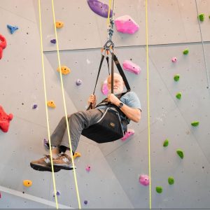 A light-skinned older adult with a white beard in adaptive equipment using a climbing wall.