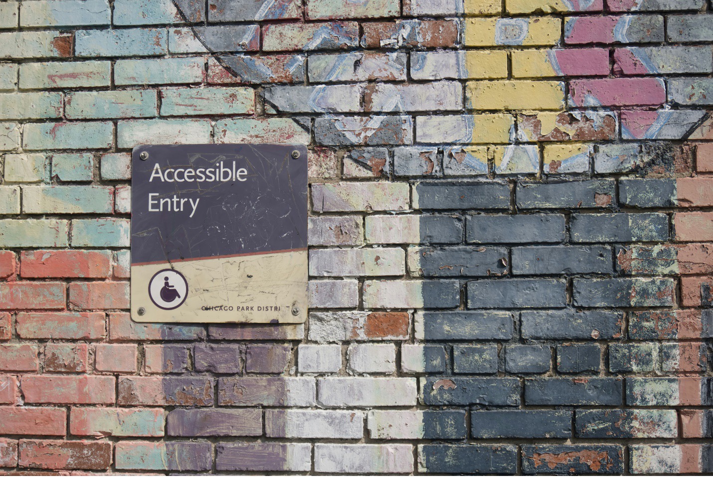 Photograph of a brick wall with a faded painted mural on it; the word "Park" can be made out. On the brick wall is a sign that reads "Accessible Entry" with a graphic of a wheelchair user on it.