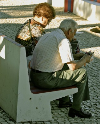 An older man and older woman sit side by side on a street bench reading.