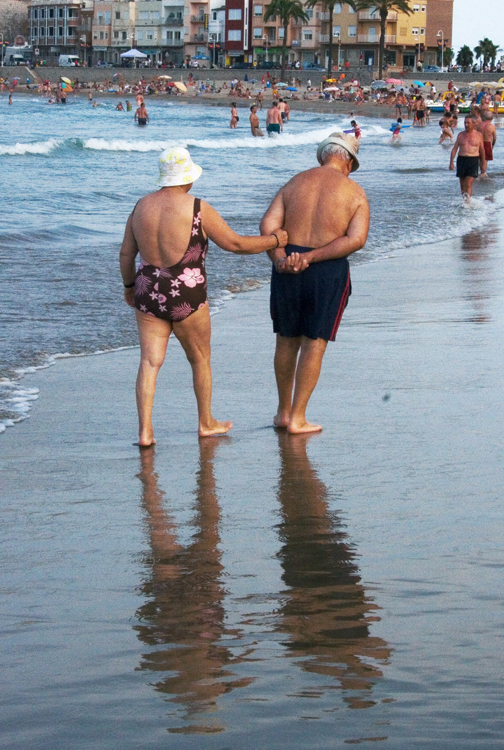 An older couple in bathing suits walk arm in arm along a beach.