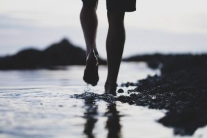 The feet and lower legs of a person walk in the water at the shore line.