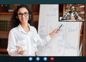 Online Tutoring. Portrait Of Woman In Glasses And Headset Having Video Conference, Teaching Foreign Languages, Talking To Camera During Web Call, Indian Student Sitting At Desk At Home And Writing