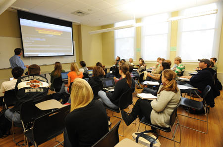 Students sitting in desks listen to the teacher at the front of the room.