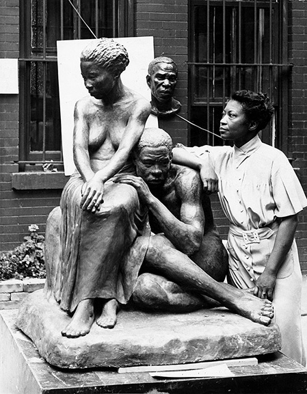 Sculptor in a dress leaning against a statue of two dark-skinned naked people