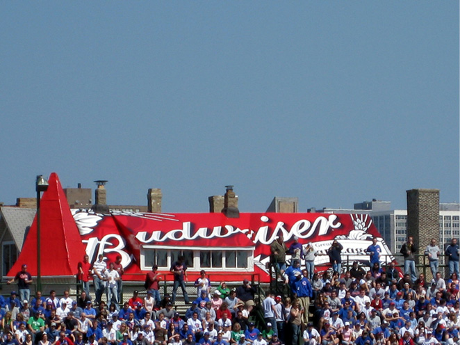A crowd of baseball fans on bleachers. The roof of the house behind them is painted with the red and white Budweiser beer logo.
