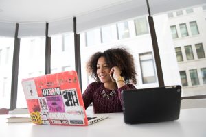 A woman of colour sits at a laptop in a corporate board room. Her laptop is covered with stickers. She is on the phone.