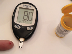 A glucometer displaying the amount of glucose measured from a finger with a yellow bottle containing the measuring strips.