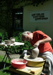 A white male in his 60s sits at a potter's wheel outside the Waterloo Potters' Workshop. He is focused on forming a vase.