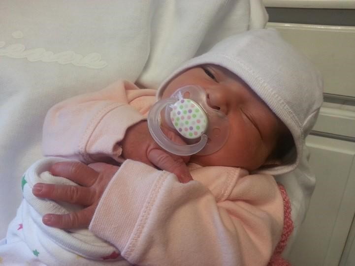 close up image of a newborn dressed in pink, wearing a white beanie hat and with a soother in her mouth