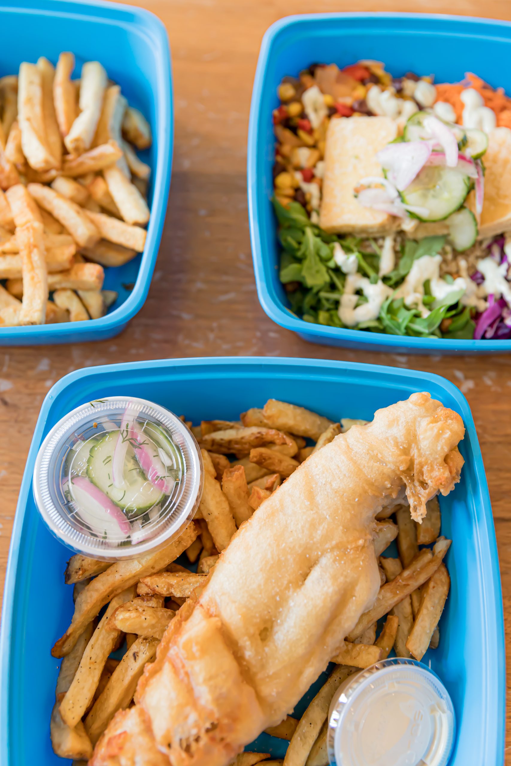Close up photo of 3 blue takeout containers with French fries, fish and chips, and salad.