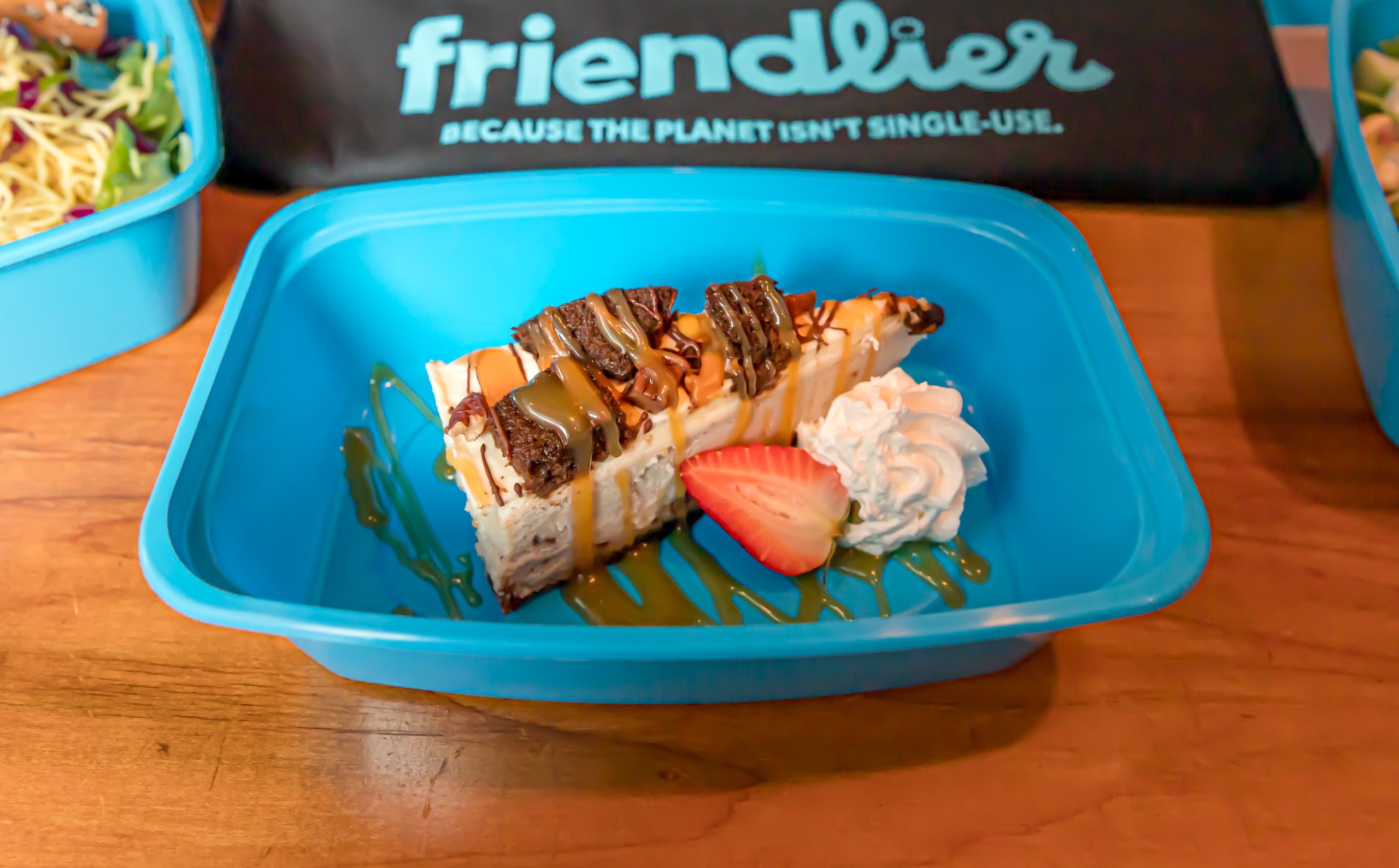 Photo of a blue takeout container with a slice of cheesecake next to Friendlier bag with motto “Because the planet isn’t single-use.”