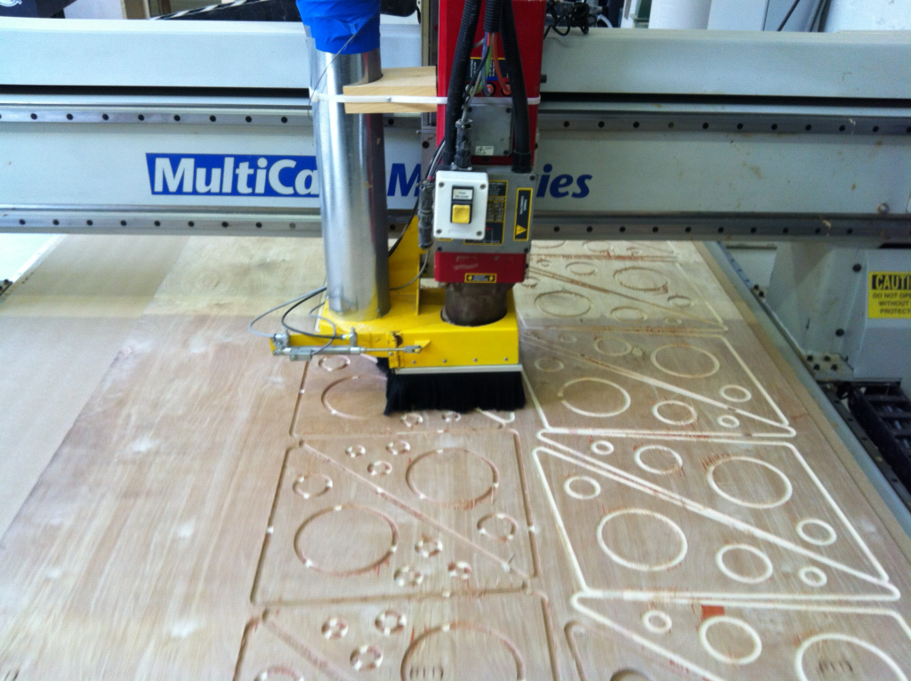 Close-up photo of the cutting arm of a CNC machine cutting a series of circles within triangles into a flat rectangular piece of sheet of wood.