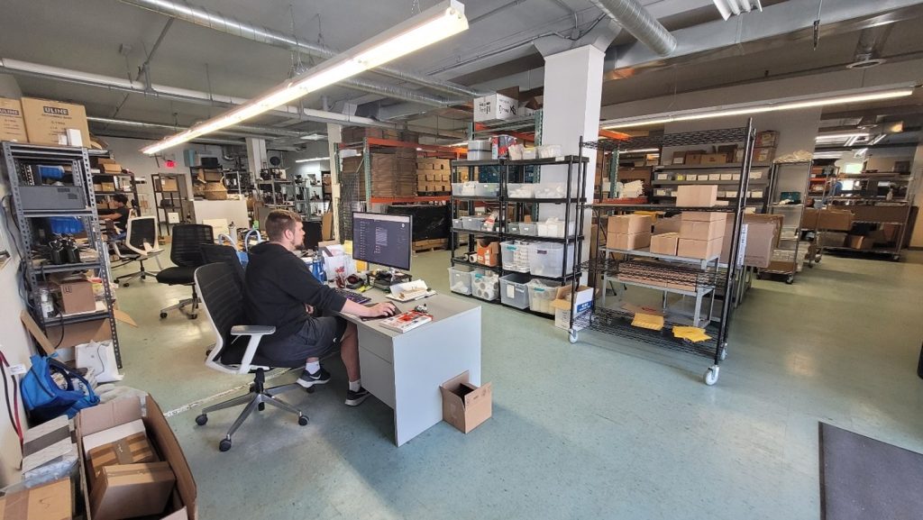 Photo of the facility with man sitting at computer desk in left foreground and industrial open shelves with carboard shipping boxes in right side background.