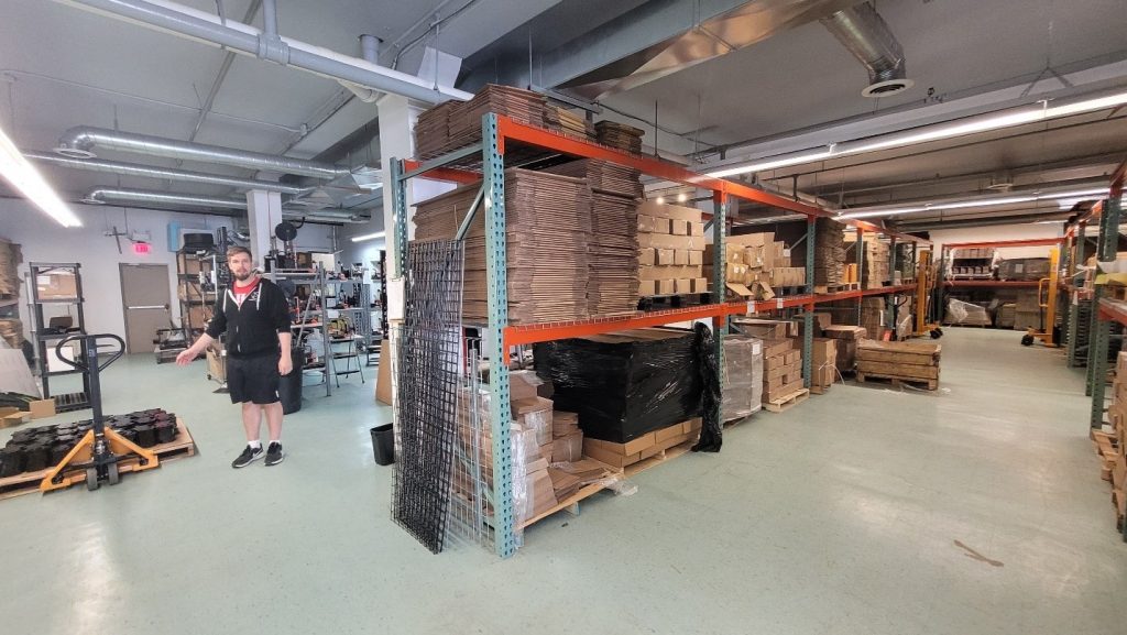 Photo of the facility with industrial open shelves at centre stacked with folded cardboard shipping boxes; assembled cardboard boxes are stacked on right of shelves; a man stands at left in background next to a pallet truck.