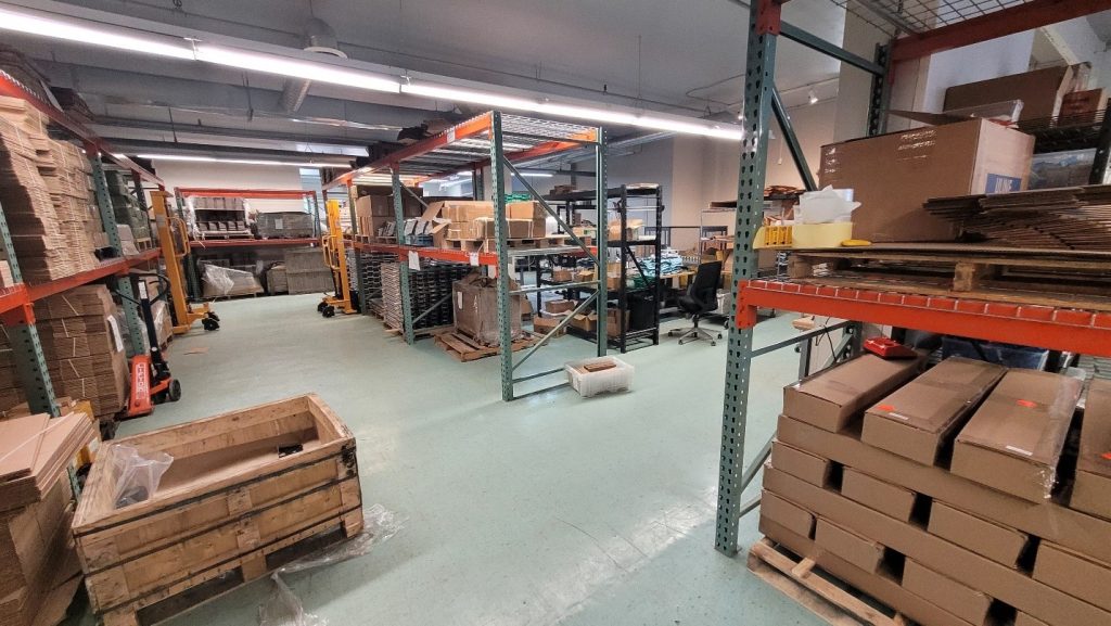 Photo of the facility with industrial open shelves of stacked boxes in right foreground; additional shelves with boxes at left and centre background. An empty pallet is in left foreground.