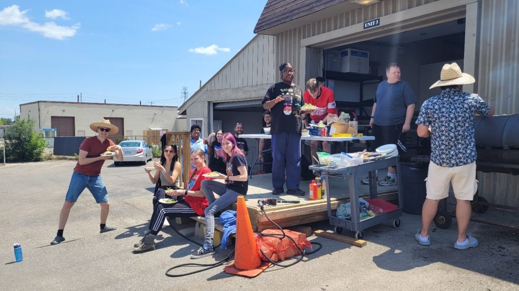 Candid photo of 13 people sitting and standing on and around the loading dock of facility on a sunny day, holding plates with food and smiling for the camera. At right a man in hat operates BBQ, at centre background is a folding project table with cook-out supplies. Orange cone and black electrical cord is in centre foreground.