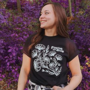 Young woman smiling outdoors in front of purple foliage, looking left, wearing a black t-shirt with white floral design and the text “Flourish and Grow.”