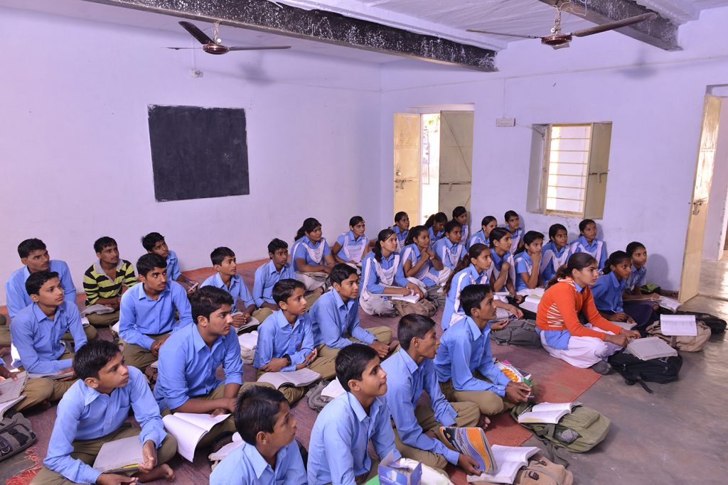 Students in blue uniforms sitting on the floor in a classroom. See image description.