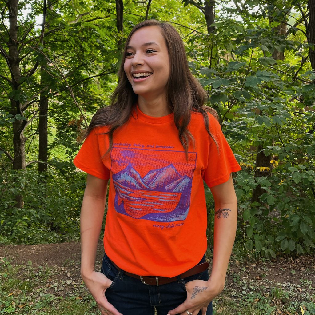 Photo of a young woman with brown hair and visible tattoos on her arm, smiling and looking left, standing in a green wooded area, wearing an orange T-shirt with a blue mountain graphic that says "yesterday, today, and tomorrow every child matters.”