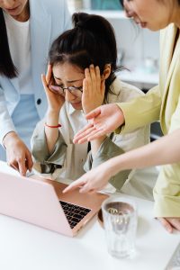 Woman sitting at computer with hands on head due to stress. Two people are trying to assist her on either side.