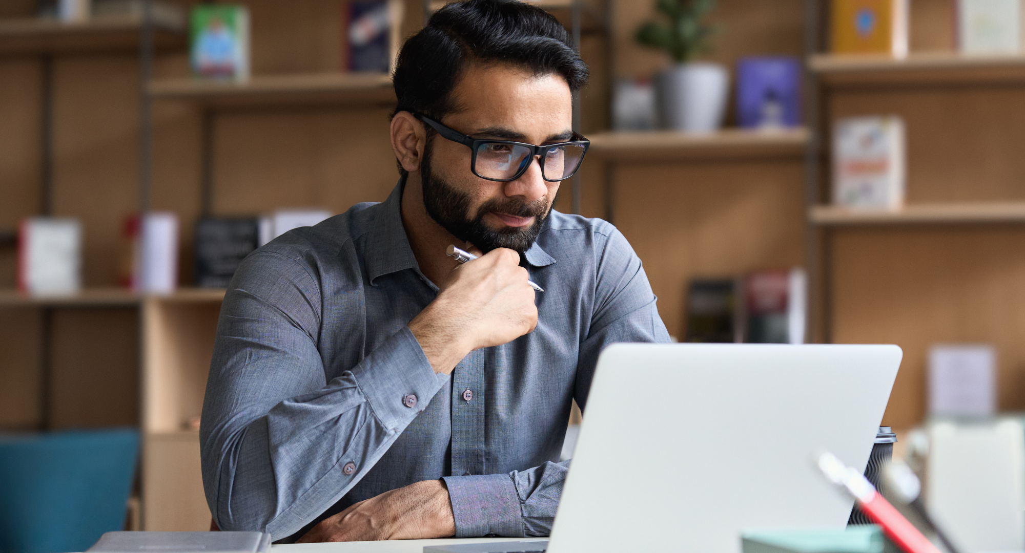 Photo of a man holding a pen and thinking to himself while looking at a laptop.