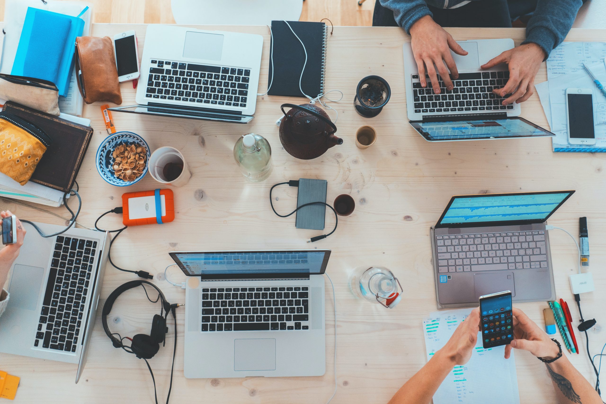 a work desk with 3 individuals behind it, 5 laptops and work-related items i.e., headphones, pens and papers together with coffee and smartphones on top.