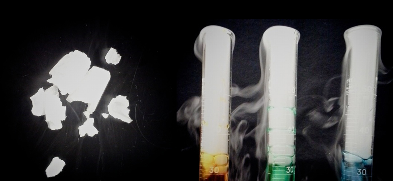A photo showing pieces of a white, sublimating substance to the left of three graduated cylinders is pictured. Each cylinder holds a different color liquid, and above the liquid, the cylinders are filled with a fog-like substance that swirls out of the top and around the outside of the cylinders.