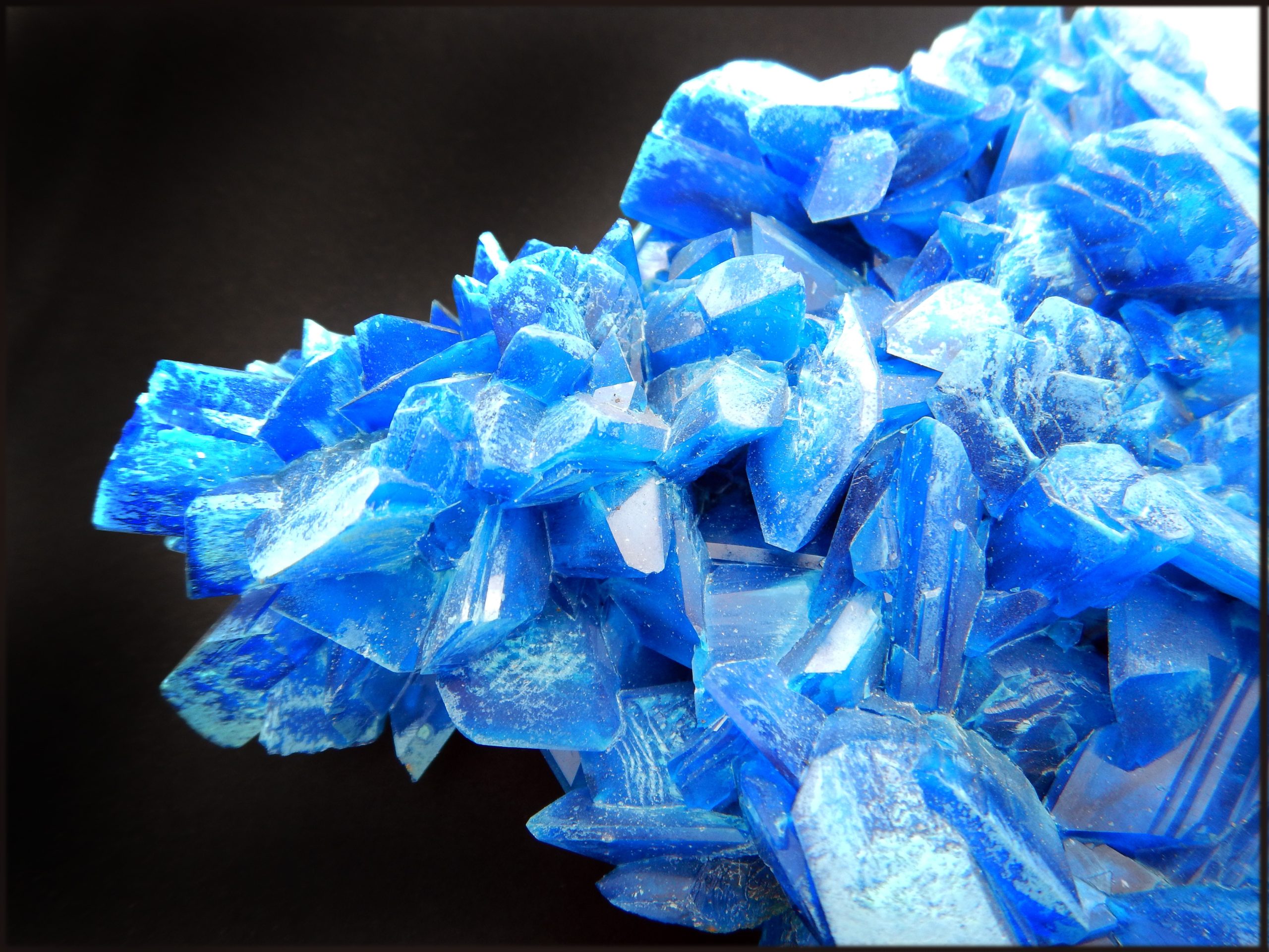 image shows royal blue solid hard crystals of copper(II) sulfate. The background colour is black.