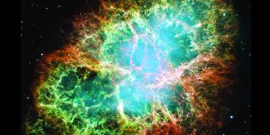 Mosaic image of the Crab Nebula in outer space showing a central green blue glow surrounded by white, gold, then burnt orange and reddish coloured filaments as you move out. Colors in the image indicate the different elements that were expelled during the explosion. White dots in the black background, surround the colourful, circular image represent stars.