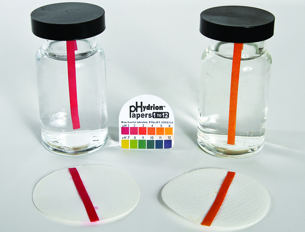This image shows two bottles containing clear colorless solutions. Each bottle contains a single p H indicator strip. The strip in the bottle on the left is red, and a similar red strip is placed on a filter paper circle in front of the bottle on surface on which the bottles are resting. Similarly, the second bottle on the right contains and orange strip and an orange strip is placed in front of it on a filter paper circle. Between the two bottles is a pack of p Hydrion papers with a p H color scale of 1 to 12 on its cover.