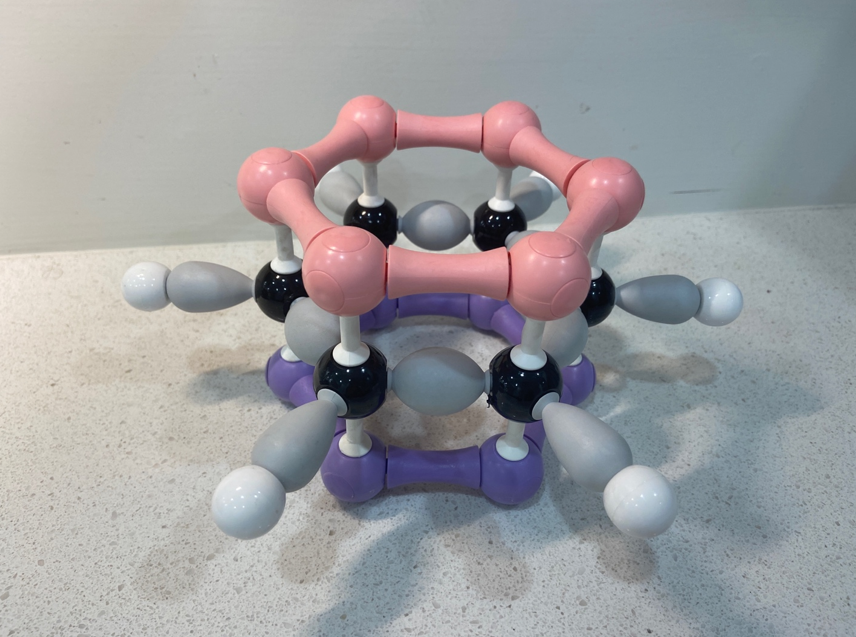 The shape of the orbitals in benzene. The top portion is pink (like the top half of a hamburger bun or bread in a sandwich), the bottom portion is purple (like the bottom half of a hamburger bun or bread in a sandwich), and the middle part shows the carbon atoms in black, hydrogen atoms in white, and all the bonds between them in grey.