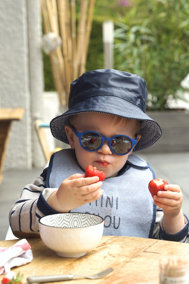 A baby wearing sunglasses and a hat eating a strawberry.