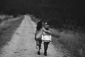 Two children walking along a path. The child on the left has their arm around the child on the right. The picture is in black and white.