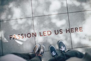 Two people standing on a sidewalk, we can only see their feet and legs. Spray painted on the sidewalk is the phrase "passion led us here."