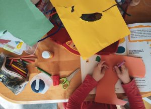 Child's craft table with yellow and green construction paper. A child is drawing on a piece of brown construction paper.