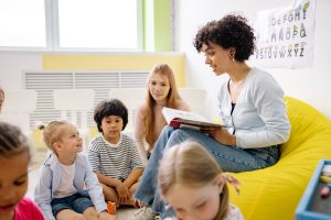 A teacher is sitting on a yellow bean bag chair while reading a book to young children.