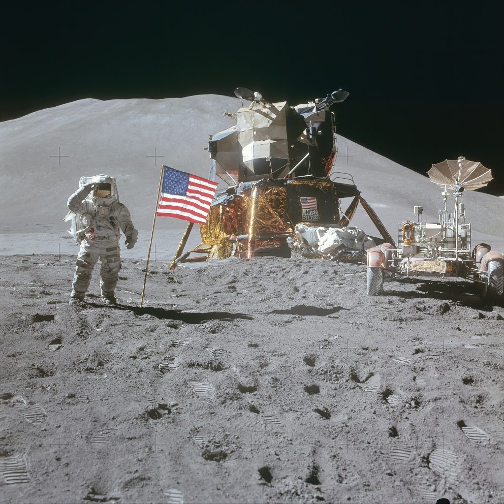 Photograph of Astronauts on the Moon. At centre is the landing module, and to the right is the Lunar rover used by the Astronauts to travel large distances from the landing site. At left an Astronaut salutes the American flag placed near the lander. Scattered throughout the foreground are footprints in the Lunar soil.