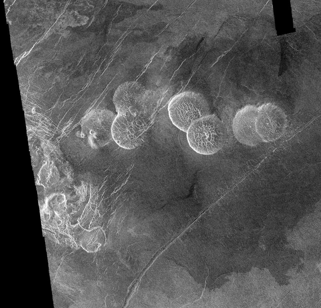 Pancake-shaped volcanoes on Venus. Five of these dome-shaped volcanoes are shown in this image. Three are clustered together in the centre left of the photograph, and two others are clustered together in the centre right of the photograph.