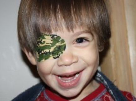 A young child smiling and wearing a green eye patch.