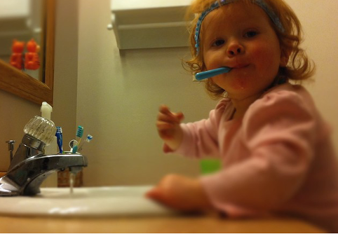 Little girl wearing headband standing in front of the sink with a toothbrush in her mouth.