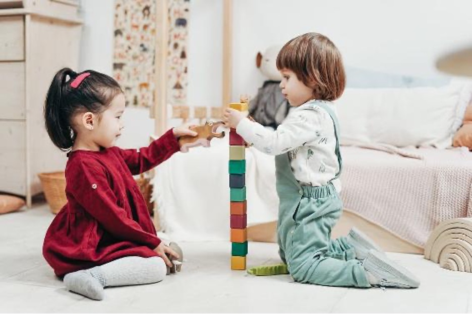 A young boy and girl kneel together on the floor as they build a block tower.