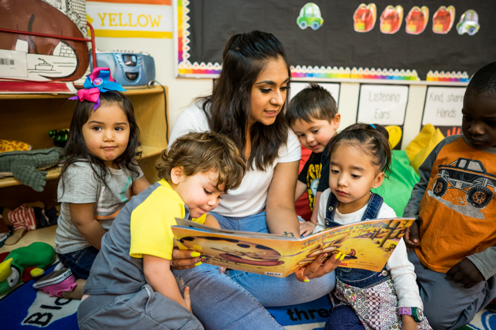 A teacher sits in the middle of a group of children in a classroom and is reading a book to them.
