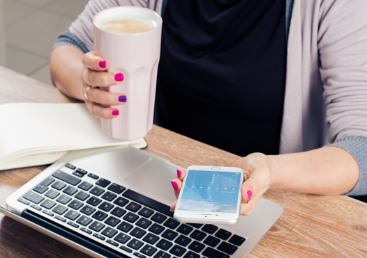 A person sits in front of a laptop holding a coffee in one hand and a phone in the other.
