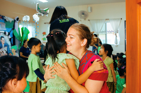 A woman hugs a child in a classroom where other children and adults are present.